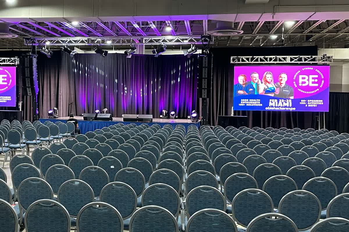 An LED screen on truss beside a draped stage in a large convention hall, showing a purple event graphic, with rows of empty banquet chairs facing the stage.