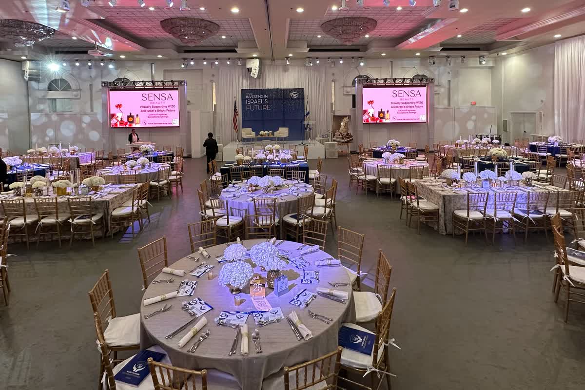 A gala ballroom set with round tables, an LED screen on either side of a draped stage showing the same sponsor graphic.