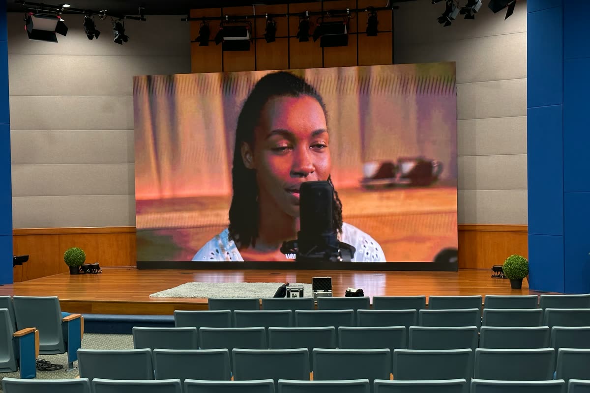 An LED wall on a low wooden stage in a corporate auditorium, playing video of a woman speaking into a microphone, with empty seats in front.
