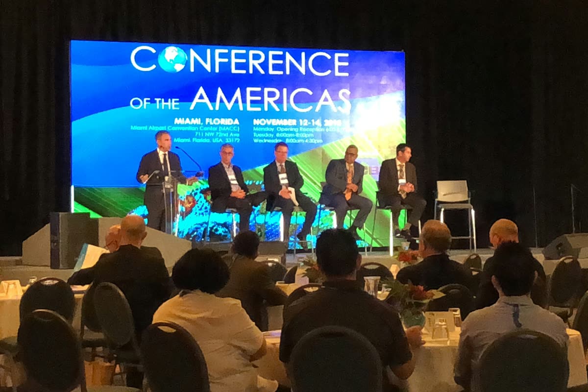 A five-person panel seated on stage in front of an LED screen reading Conference of the Americas, Miami, Florida, with an audience at banquet tables.