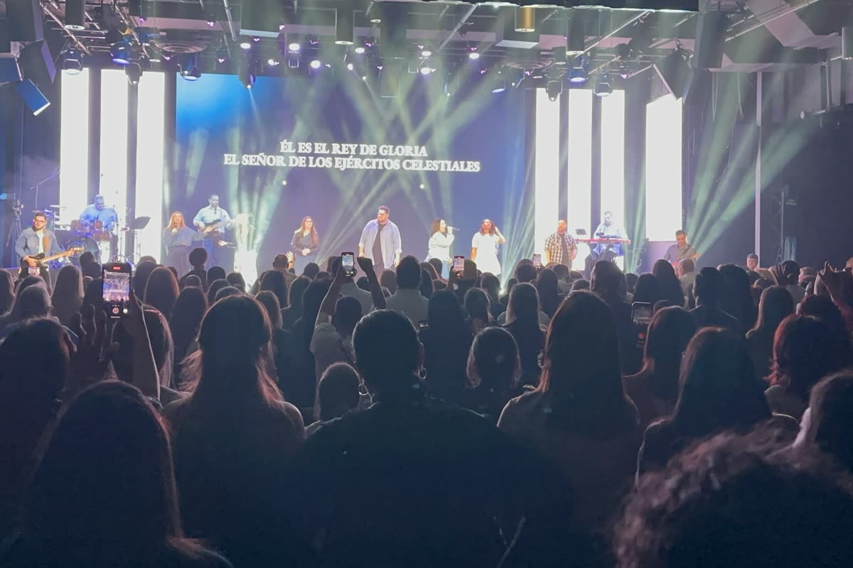 A theater stage during a live program, seen from the middle of a standing audience, the LED wall lit in white beams with lyrics across it.